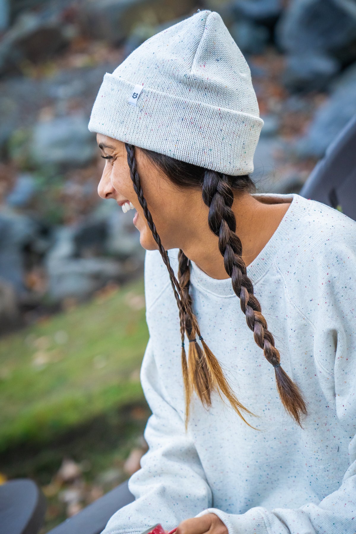 Woman wearing a gray beanie and light gray sweater with braided hair, sitting outdoors.