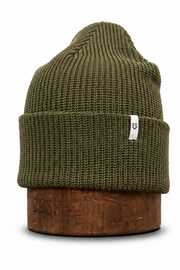 An olive green ribbed knit beanie hat displayed on a brown wooden block against a white background.