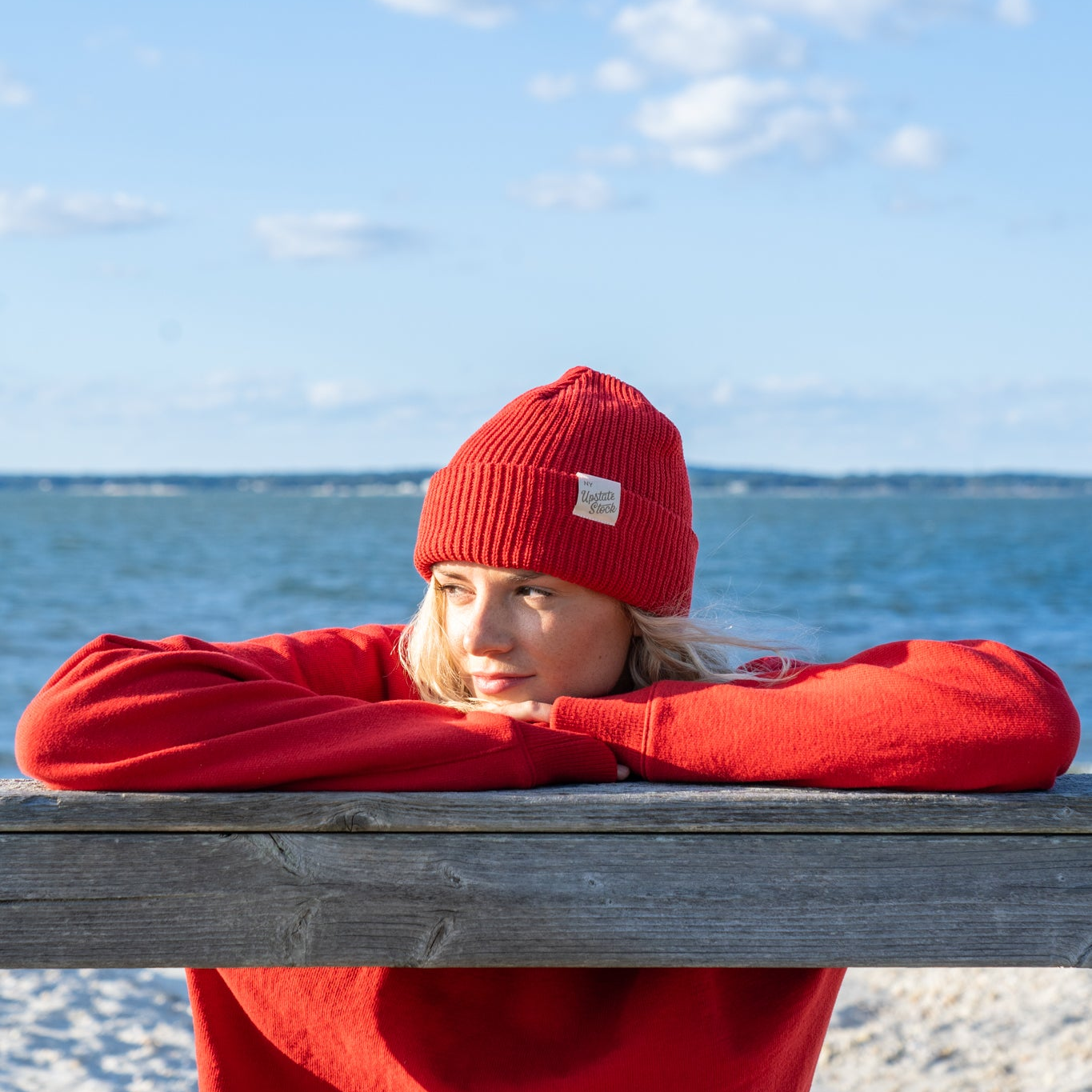 Person wearing a red beanie and sweater sitting on a wooden bench by the ocean.